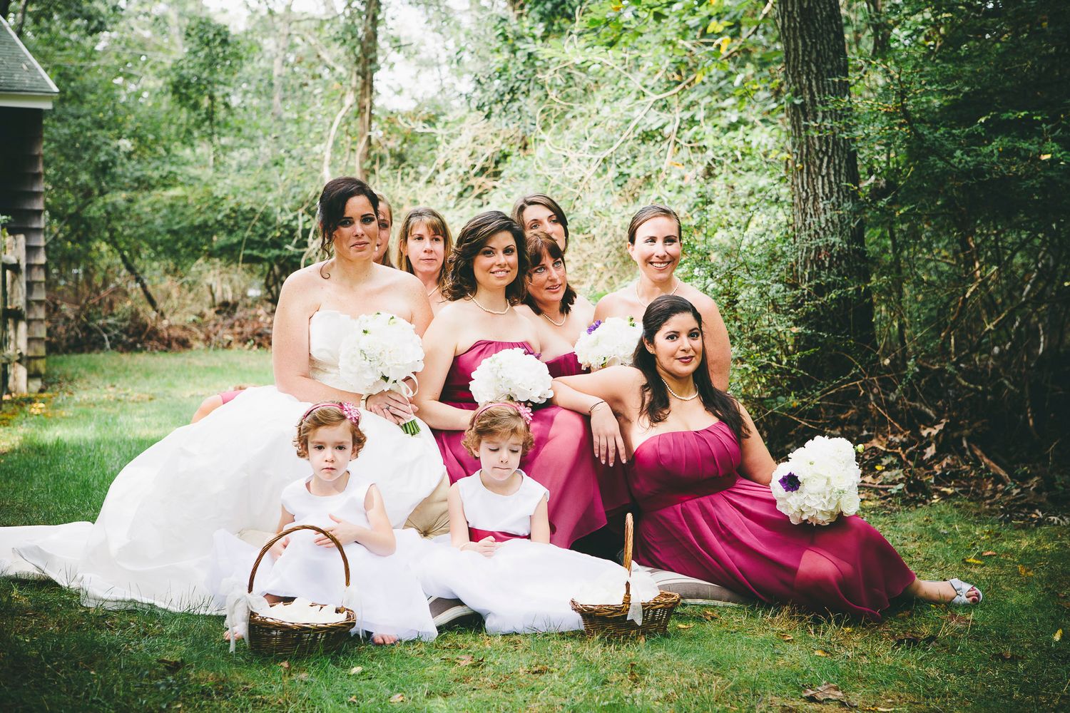 Bride and bridesmaids in burgundy dresses posing with flower girls on green lawn at outdoor wedding.