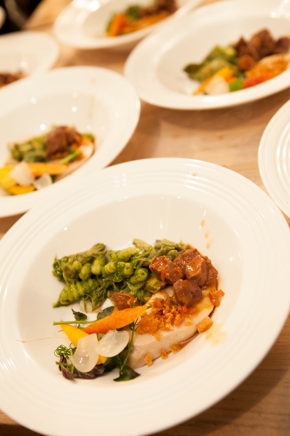 Multiple white plates of gourmet fish dish with green pea gnocchi, braised meat, and colorful vegetables for service.