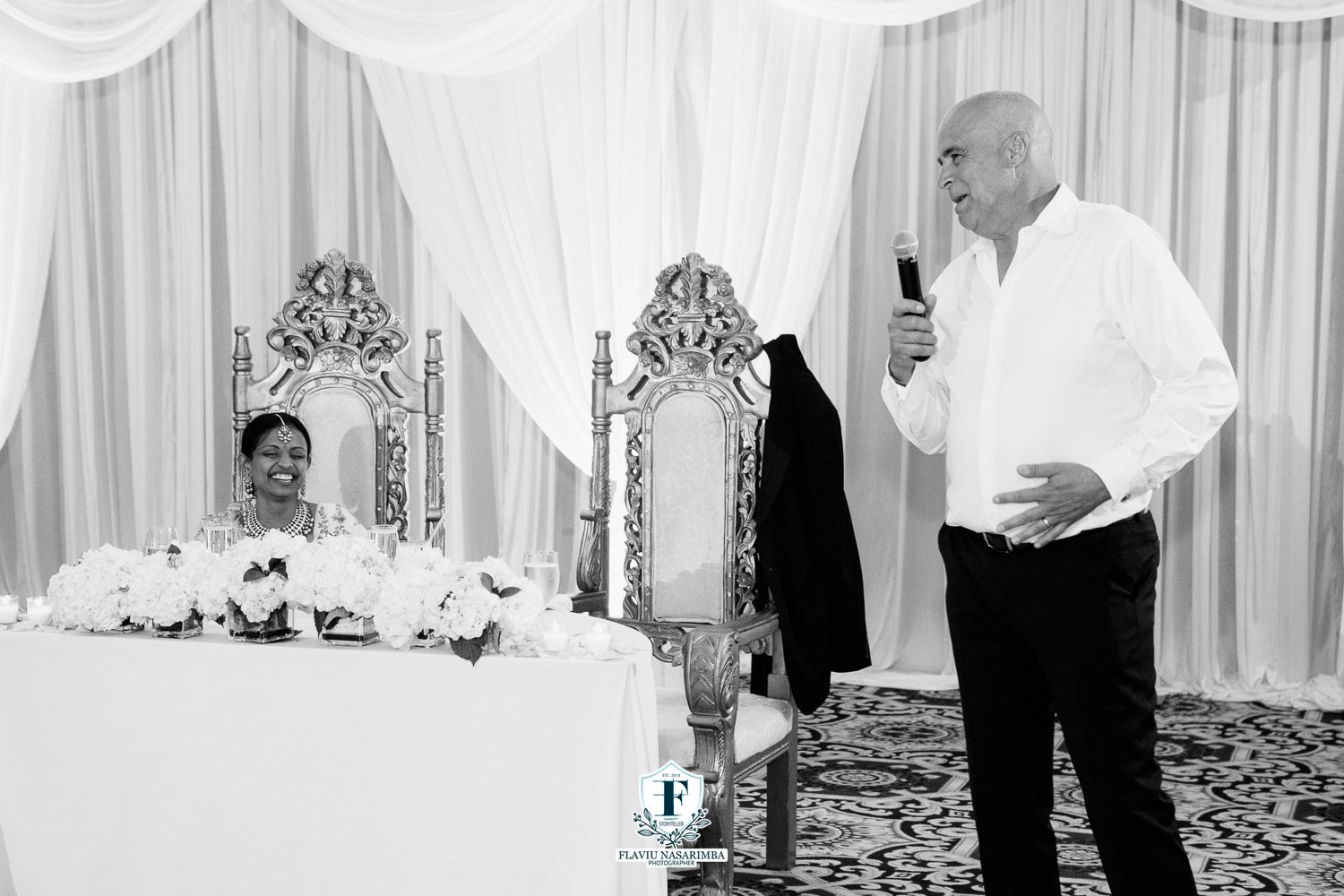 Black and white photo of groom giving speech while bride sits at floral-decorated sweetheart table at wedding.
