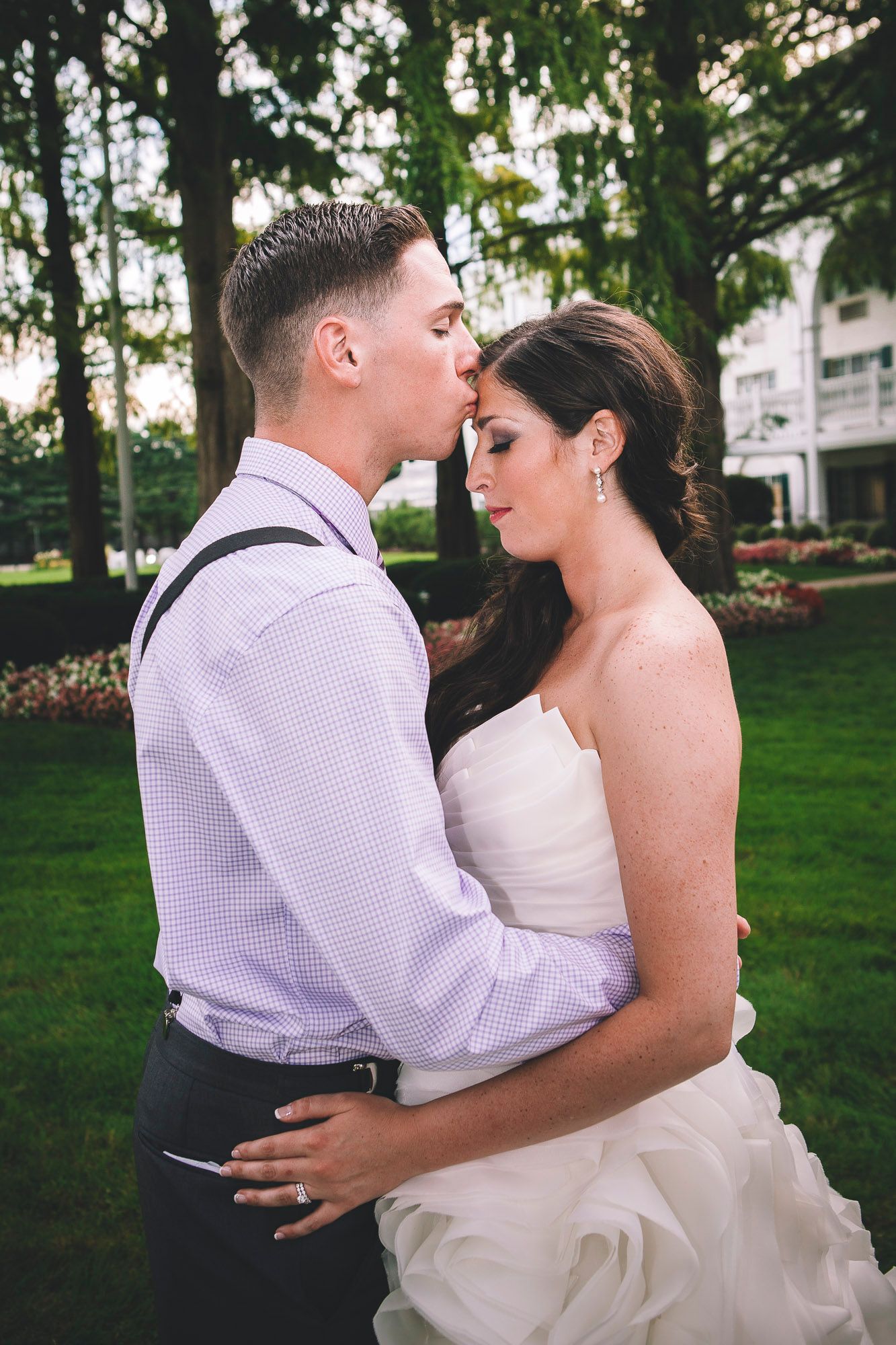 Groom tenderly kisses bride on forehead in romantic outdoor wedding portrait on lush green lawn.