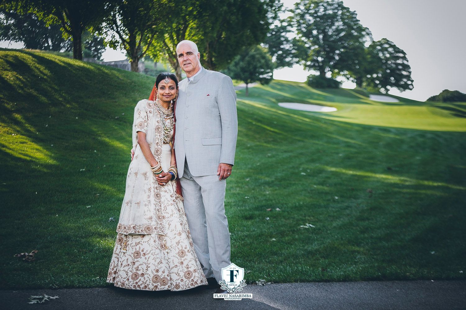 Interracial newlywed couple posing on golf course, bride in gold lehenga and groom in grey suit.