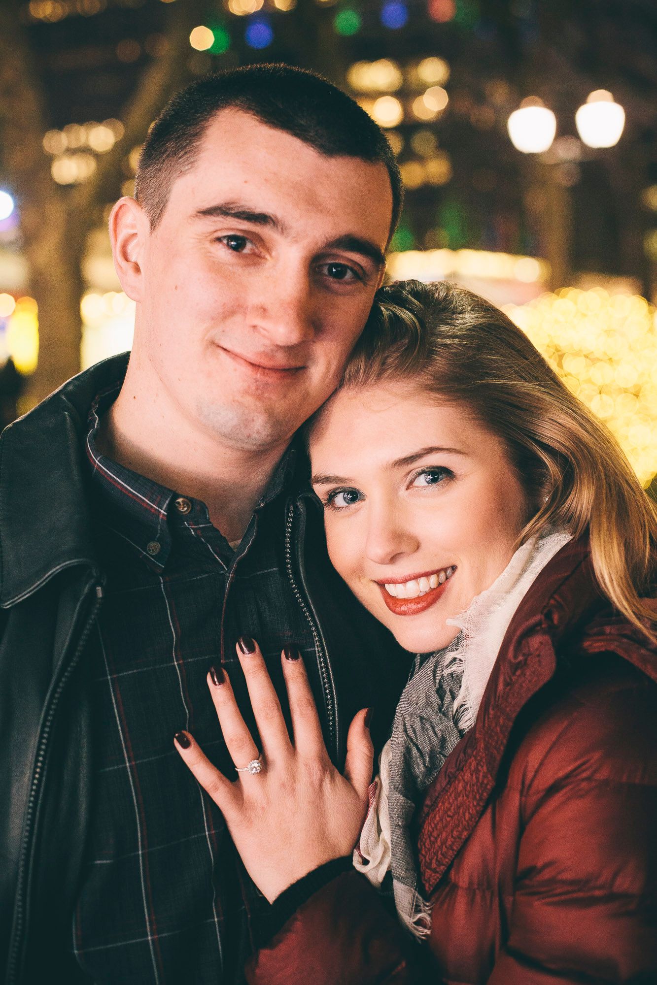 Happy couple showing engagement ring with festive bokeh lights at night.