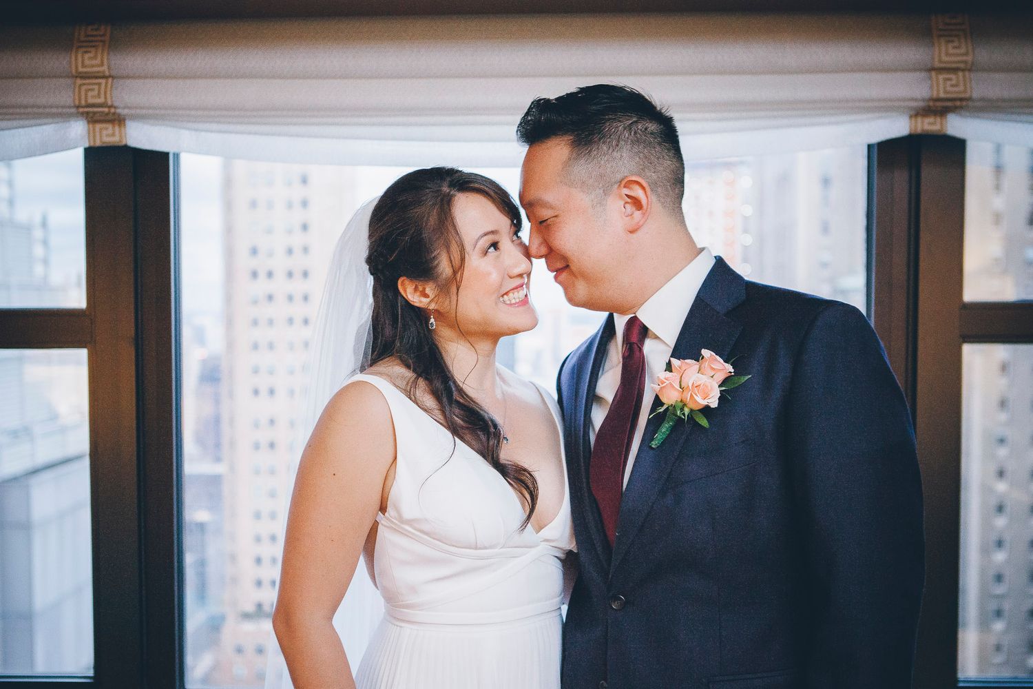 Bride and groom share a tender moment touching foreheads by a window with city skyline views.