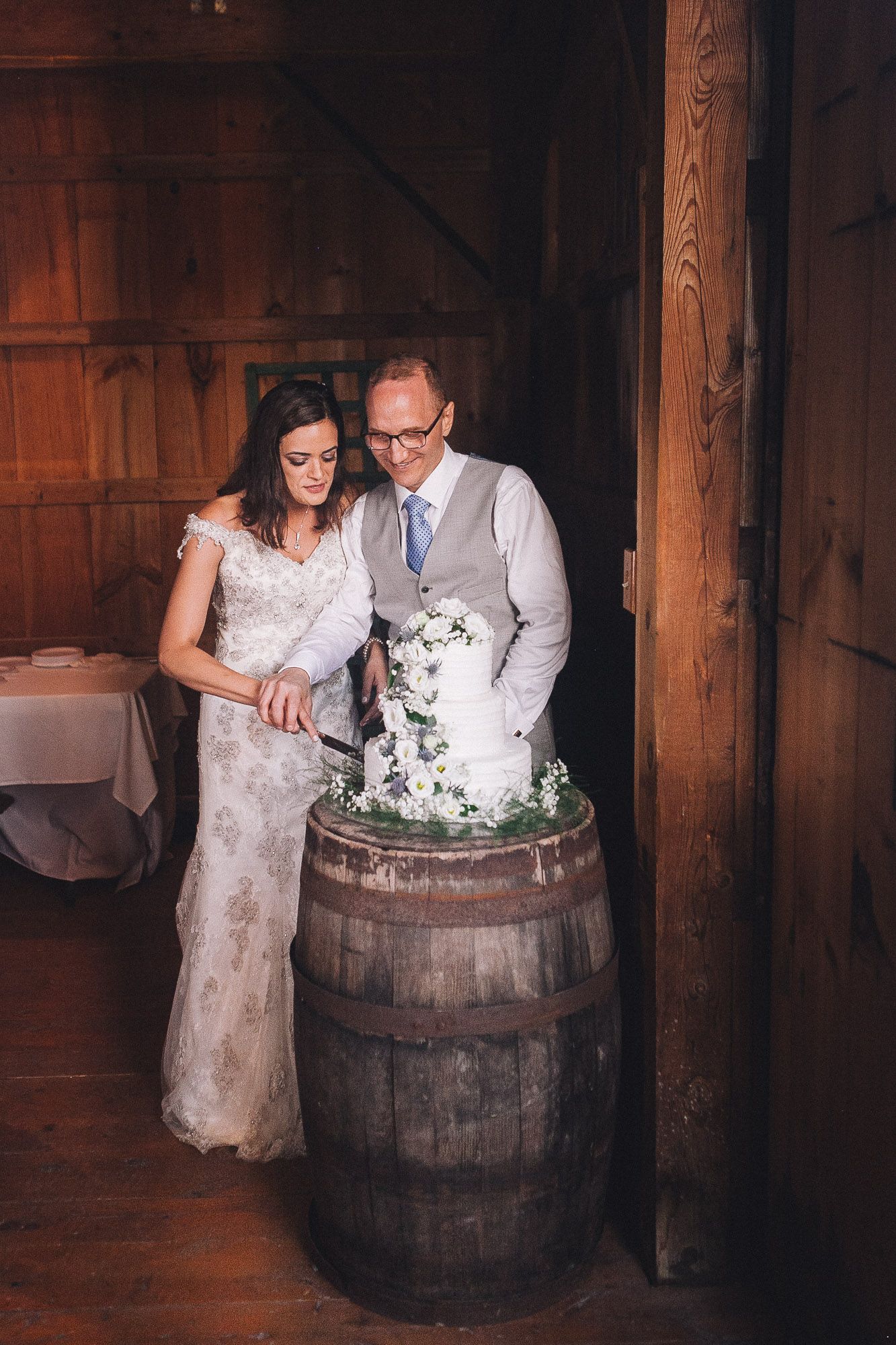 Bride and groom cut their white floral wedding cake displayed on a rustic wooden barrel.