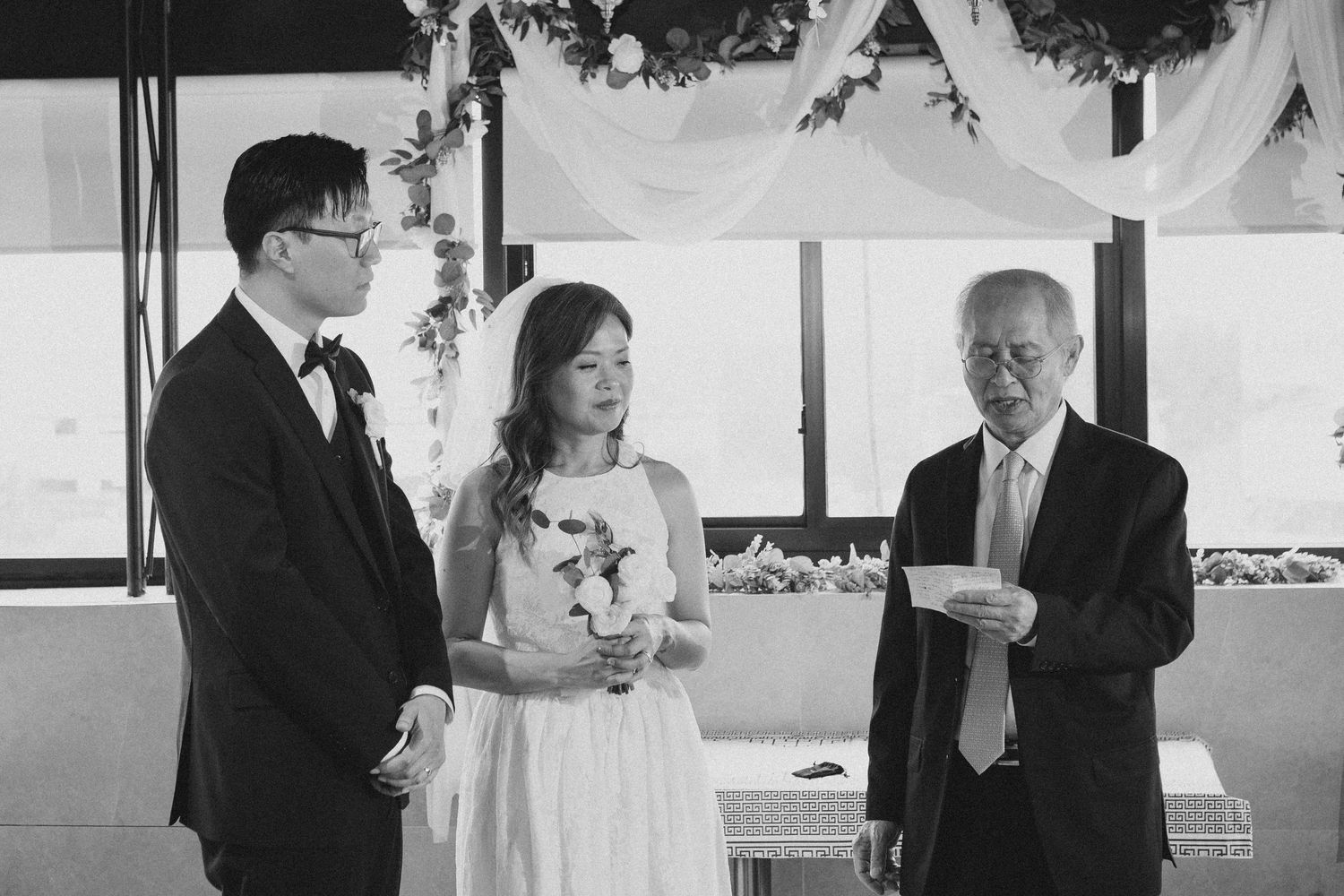 Black and white photo of bride and groom listening to officiant read vows at wedding ceremony.