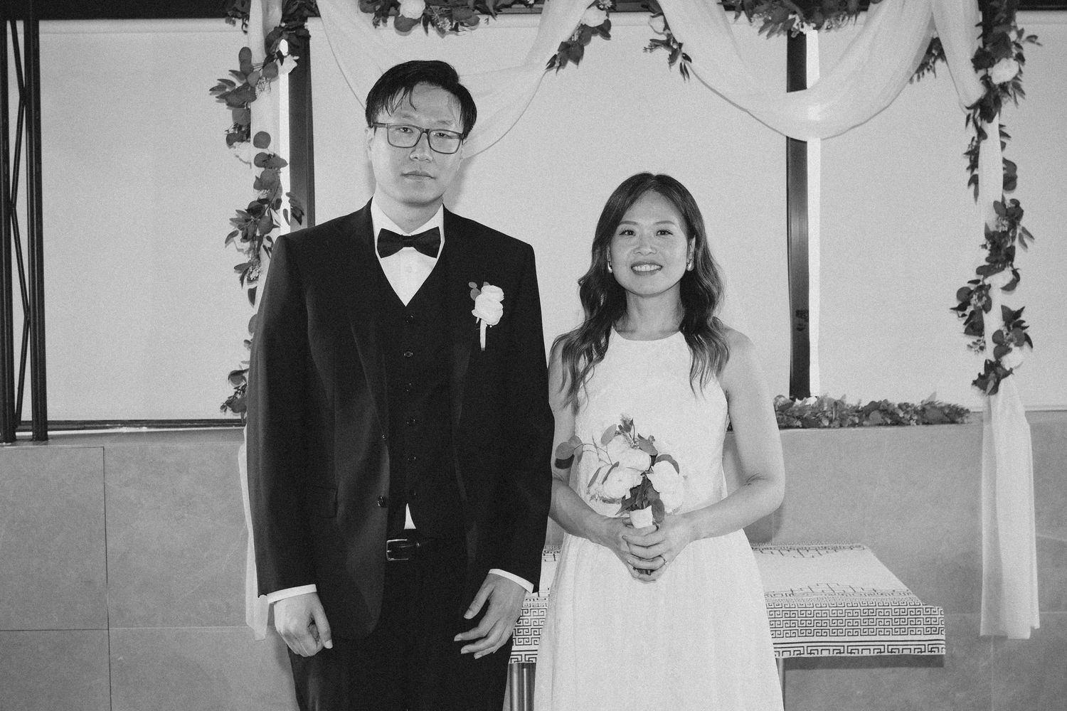 Bride and groom pose together in black and white wedding photo with floral arch backdrop.