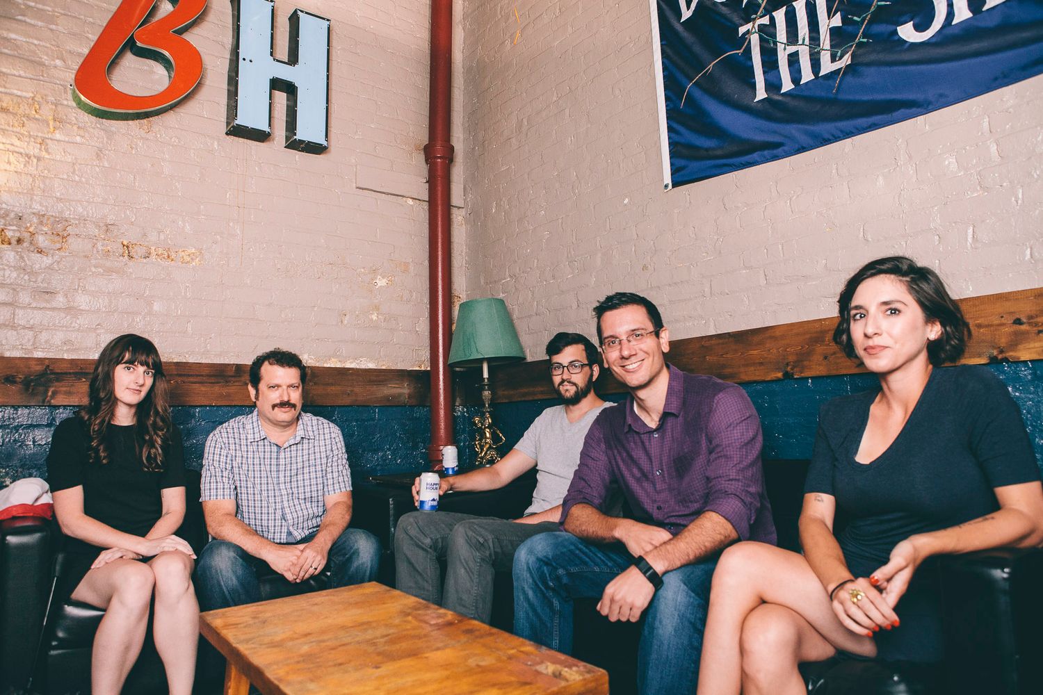 Five young professionals posing together on a couch in a hip bar with BH logo on wall.