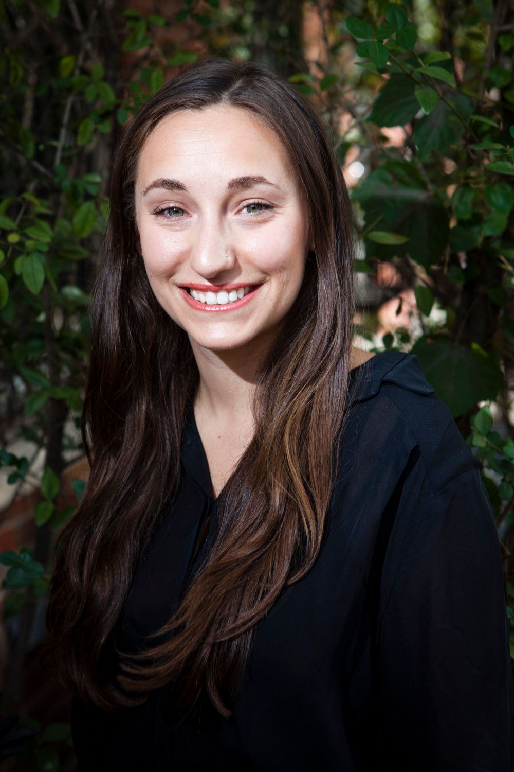 Young woman with long brown hair smiling outdoors wearing a black blouse.