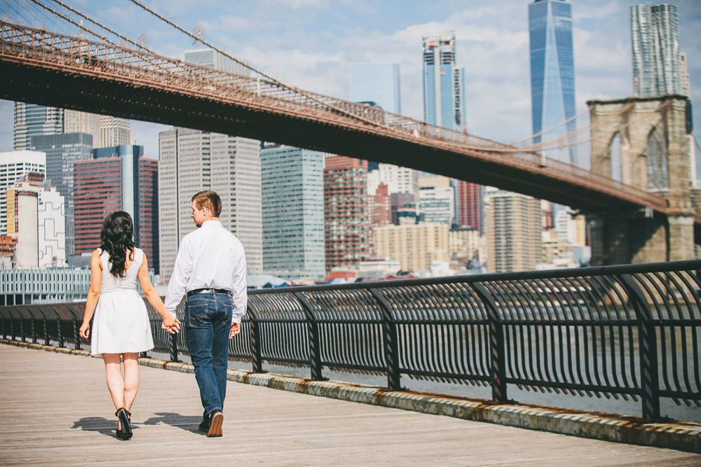 Couple holding hands walking along Brooklyn Bridge promenade with NYC skyline in background.