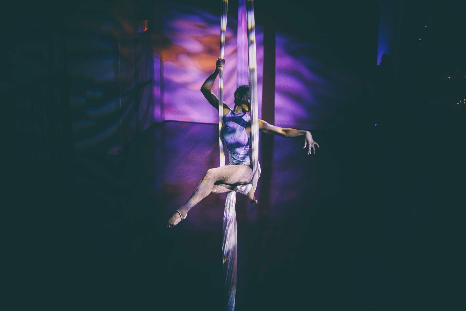 An aerial silk performer executes a graceful split pose in a dimly lit purple-hued event space.