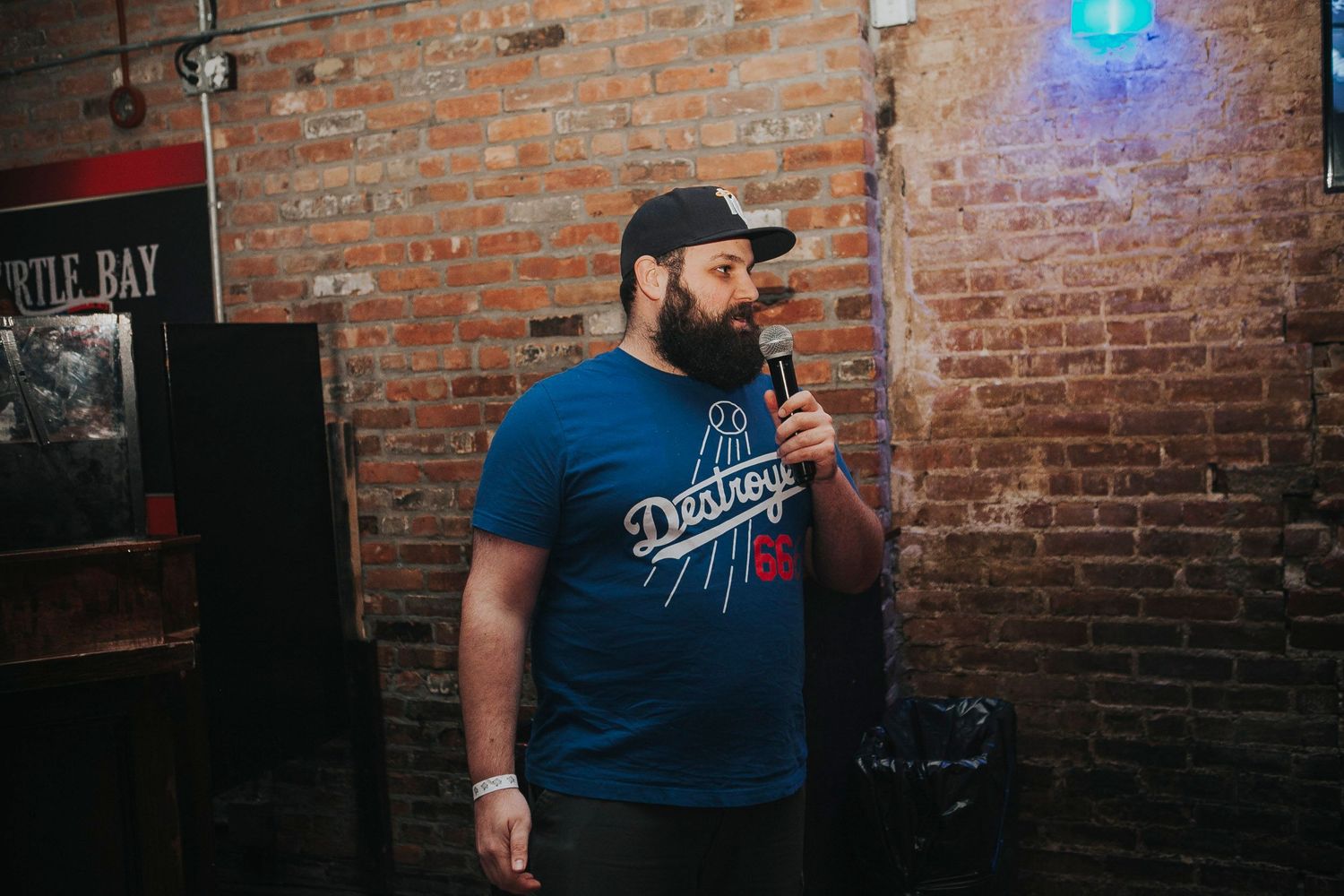 Bearded man in a blue Dodgers t-shirt speaking into a microphone at a brick-walled venue.