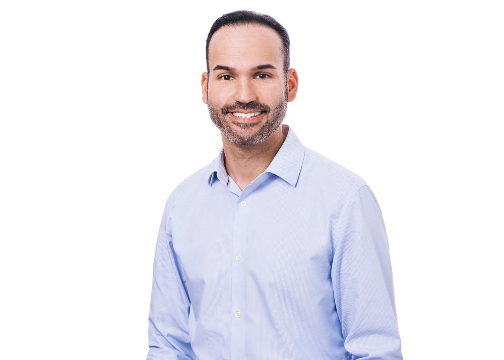 Professional headshot of a smiling bearded man in a light blue shirt on a white background.