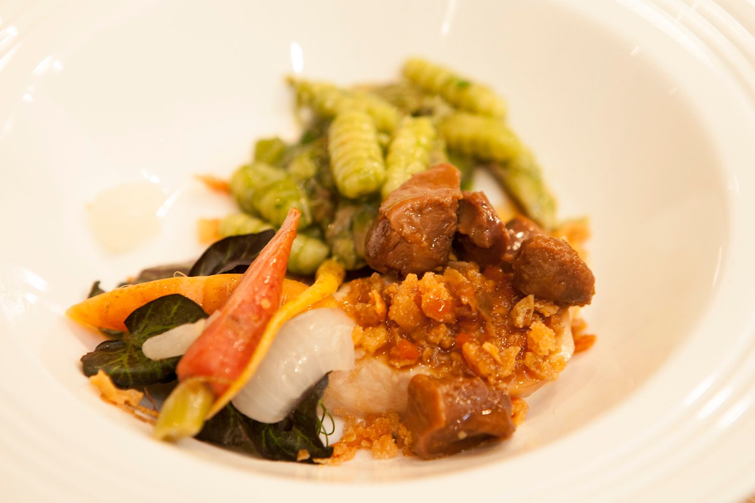 Fine dining dish featuring white fish, green pea gnocchi, braised meat, sauteed greens, and carrots in a white bowl.