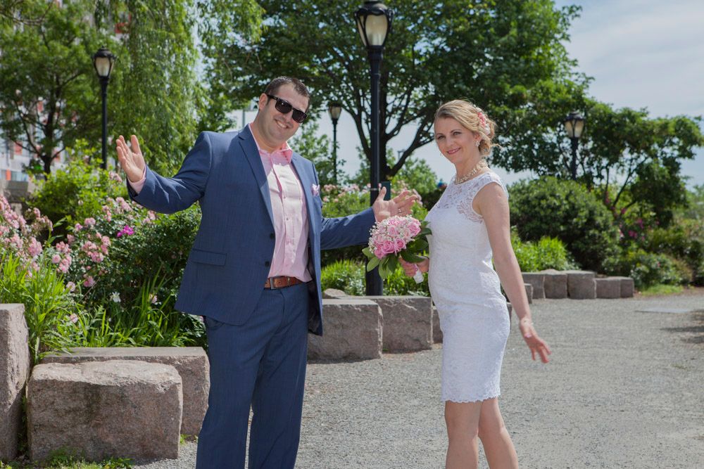 Groom in navy suit waves while bride in white dress holds pink bouquet in a sunny garden setting.