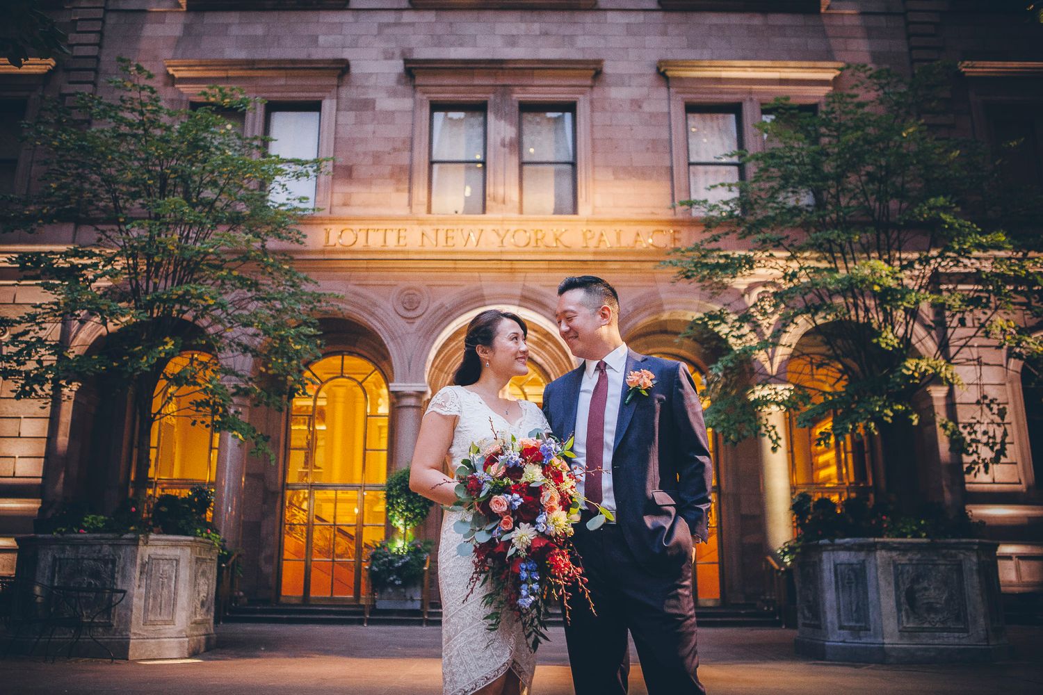 Newlyweds share a romantic look outside an elegant illuminated building at dusk.