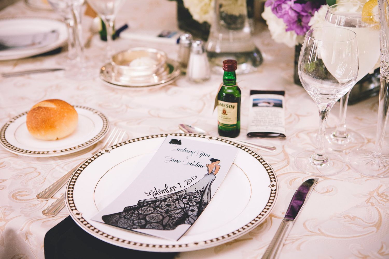 Elegant wedding reception place setting with illustrated menu card, bread roll, and mini whiskey bottle.