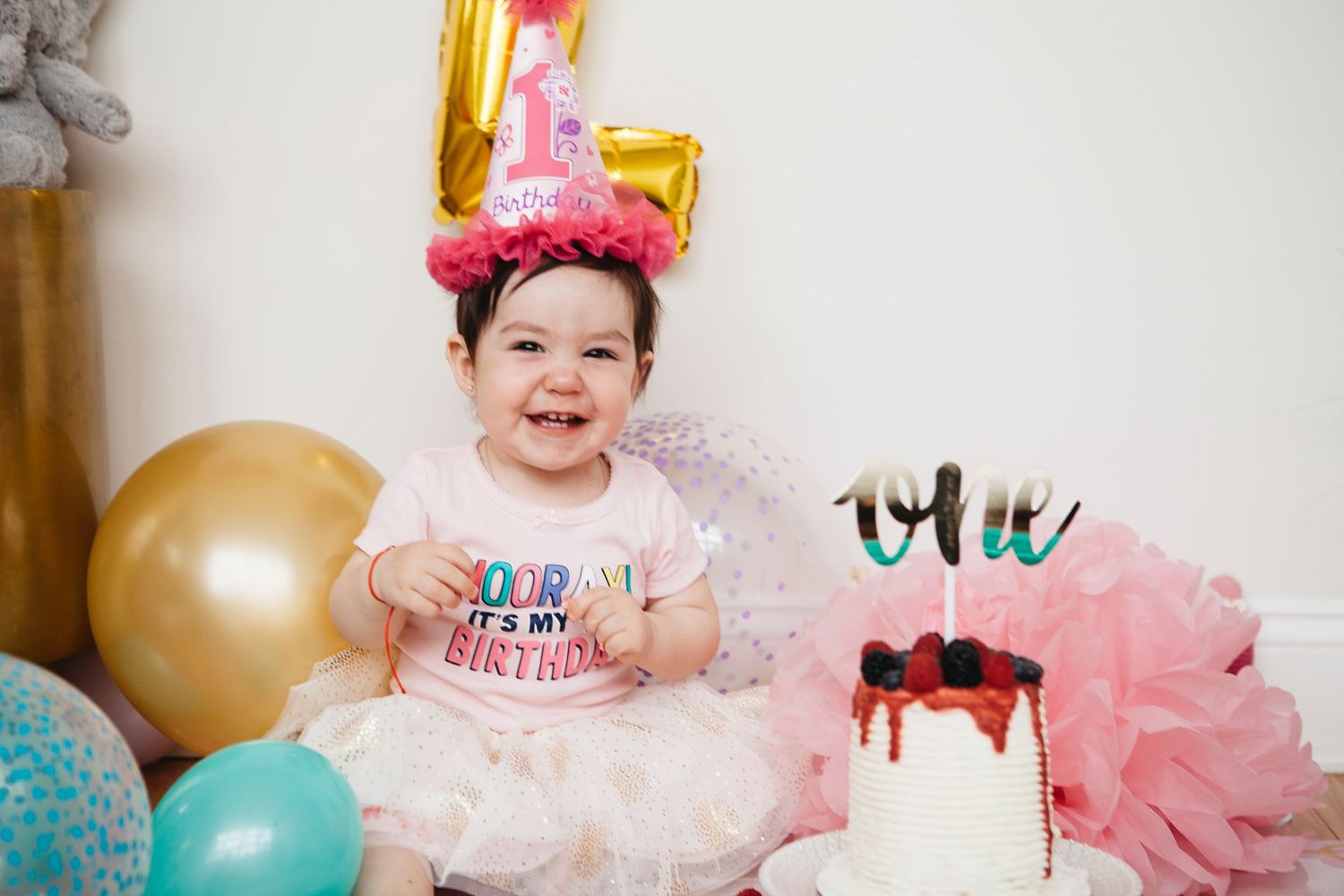 Happy baby girl in pink tutu and birthday hat smiles beside her first birthday cake with balloon decor.