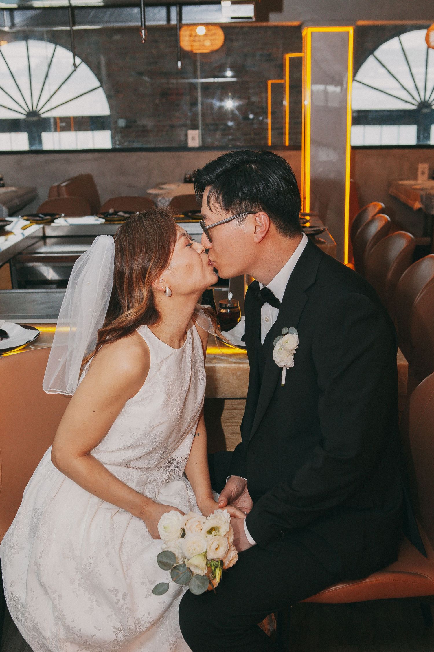 Asian bride and groom share a romantic kiss at wedding reception holding white floral bouquet.