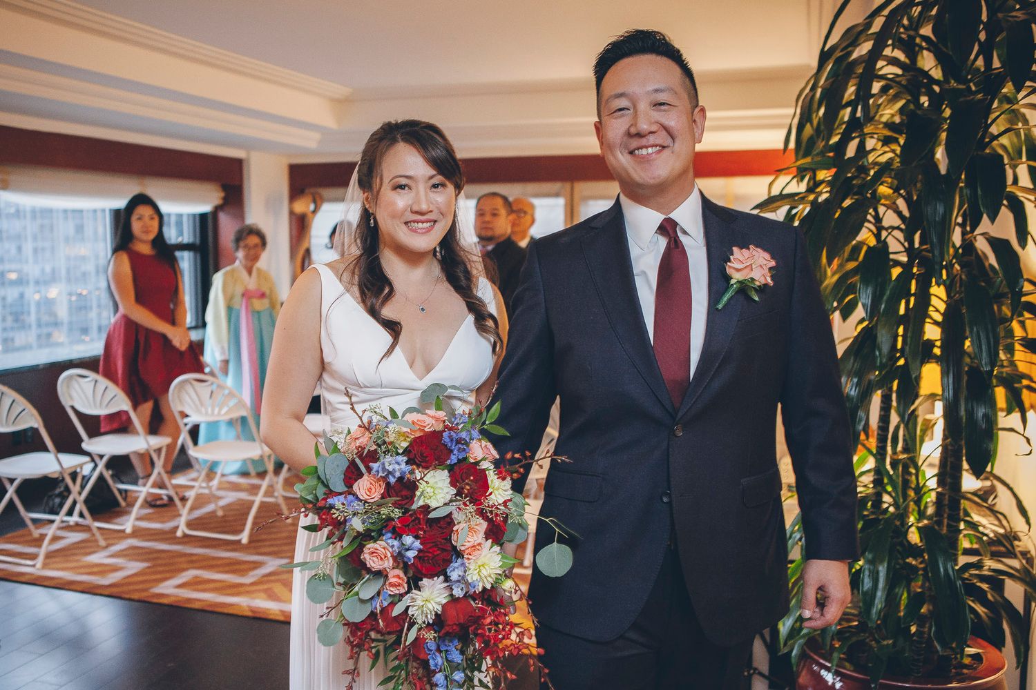 Smiling newlyweds walk together indoors, bride holding a lush red and white bouquet.