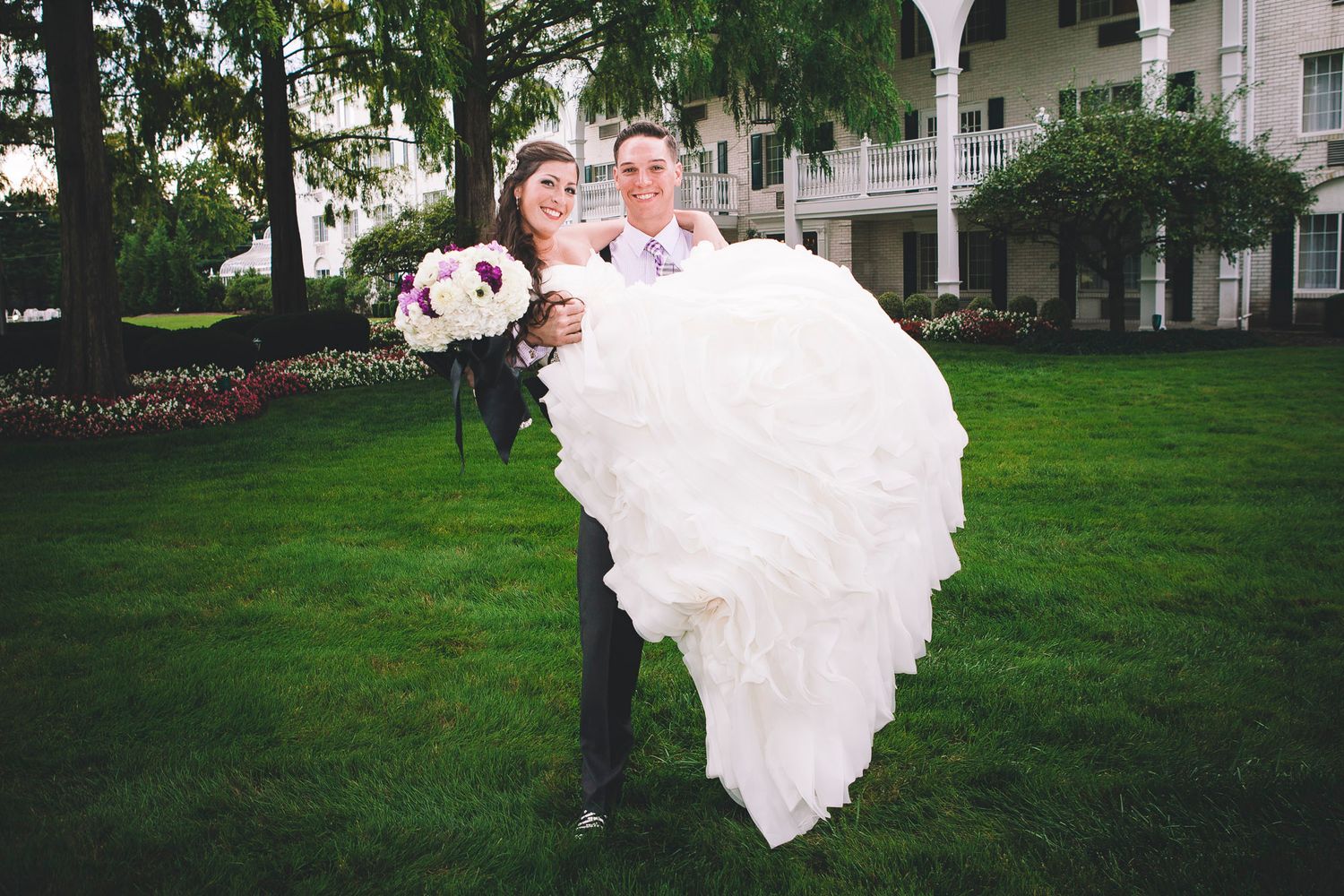Smiling groom carries bride in voluminous white wedding gown on lush green lawn.