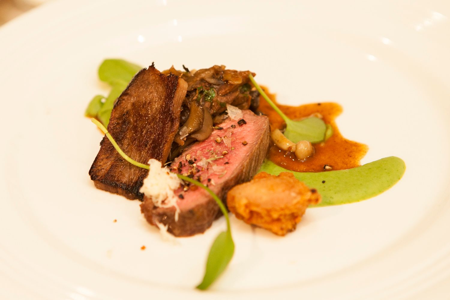 Gourmet plated lamb dish with pink sliced meat, green pea puree, mushrooms, and rich sauce on a white plate.