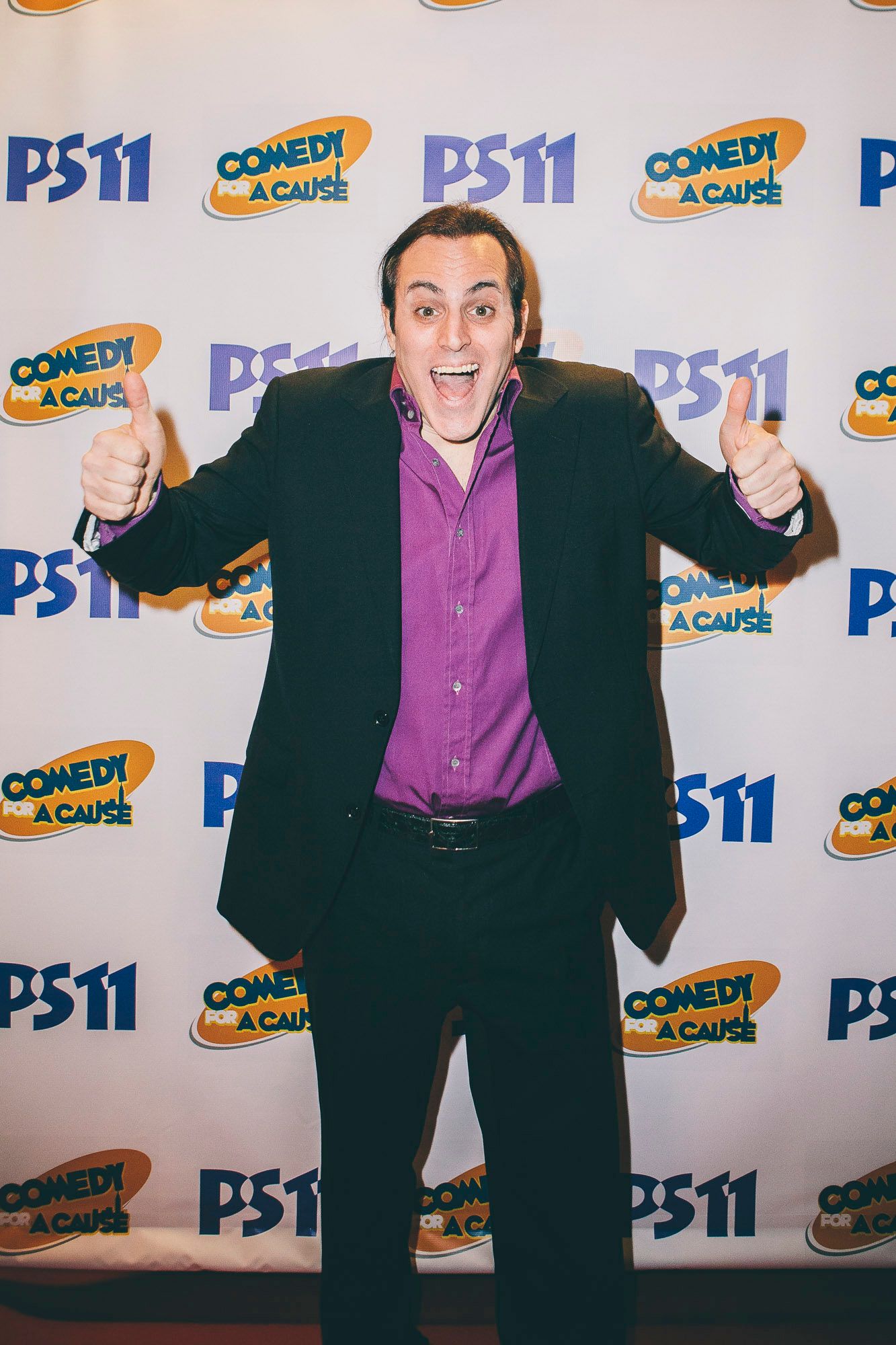Enthusiastic man in purple shirt giving thumbs up at PS11 Comedy for a Cause event.