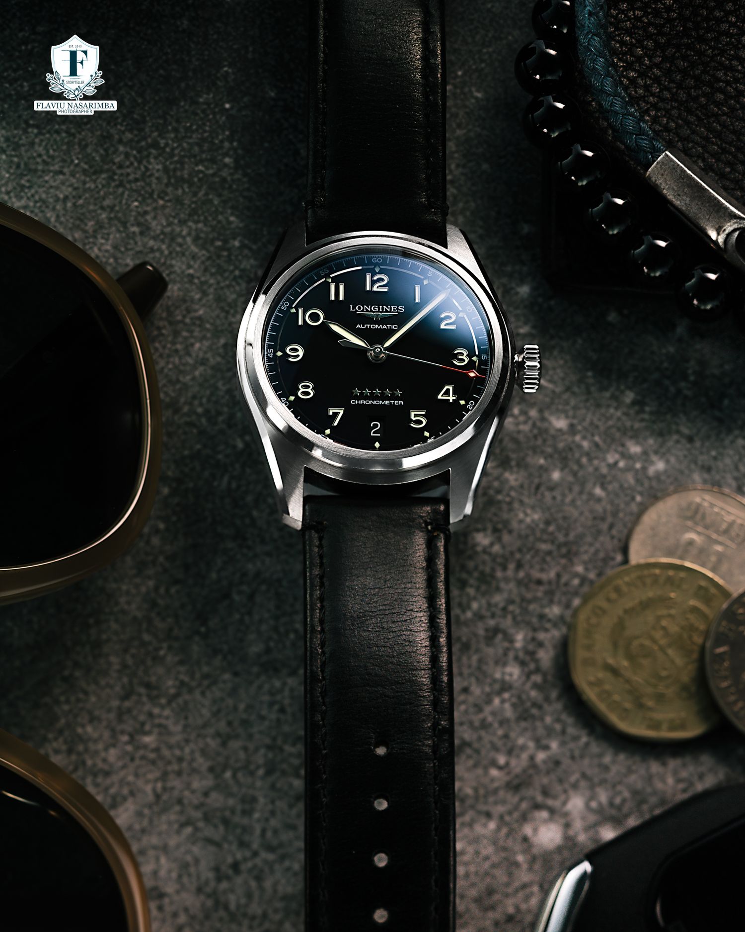 Longines black dial dress watch with leather strap displayed alongside sunglasses and coins.
