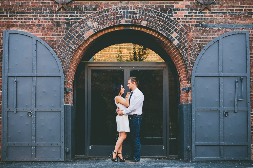 Romantic couple embracing under brick archway with large grey doors in an urban industrial setting.