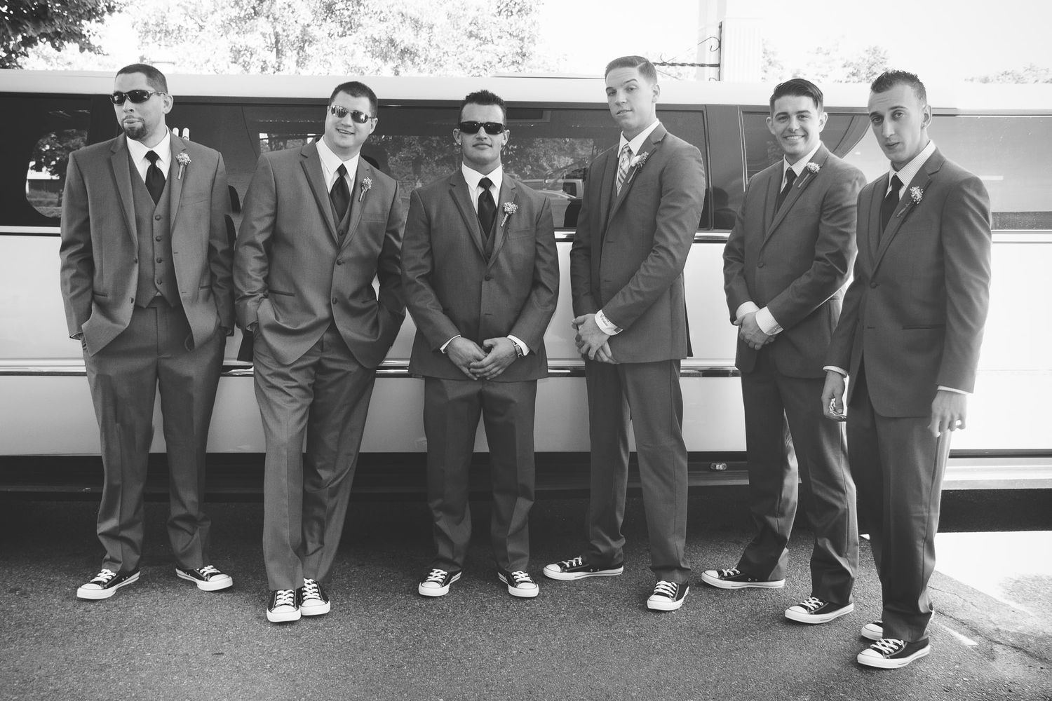 Six groomsmen in gray suits and Converse sneakers posing in front of a white limousine in black and white.