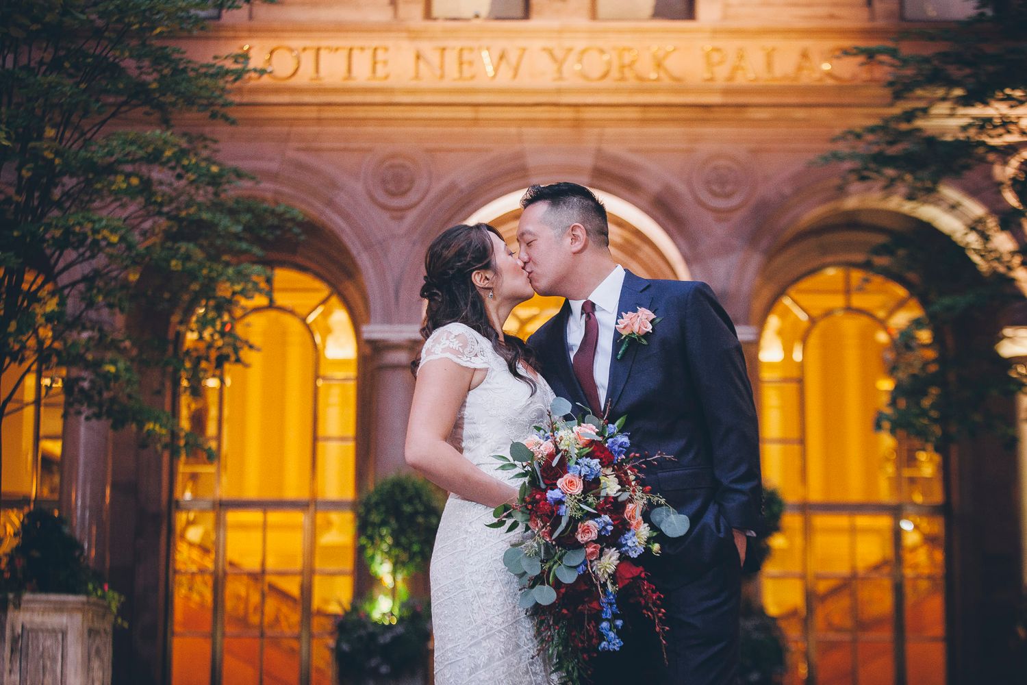 Bride and groom sharing romantic kiss outside illuminated Lotte New York Palace hotel entrance at night.