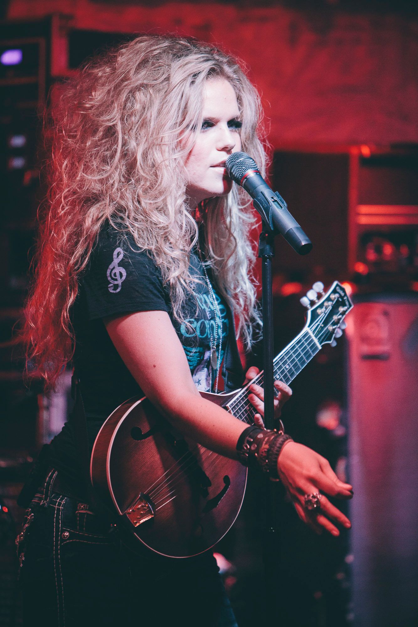 Curly blonde female musician sings into microphone while playing mandolin on stage.