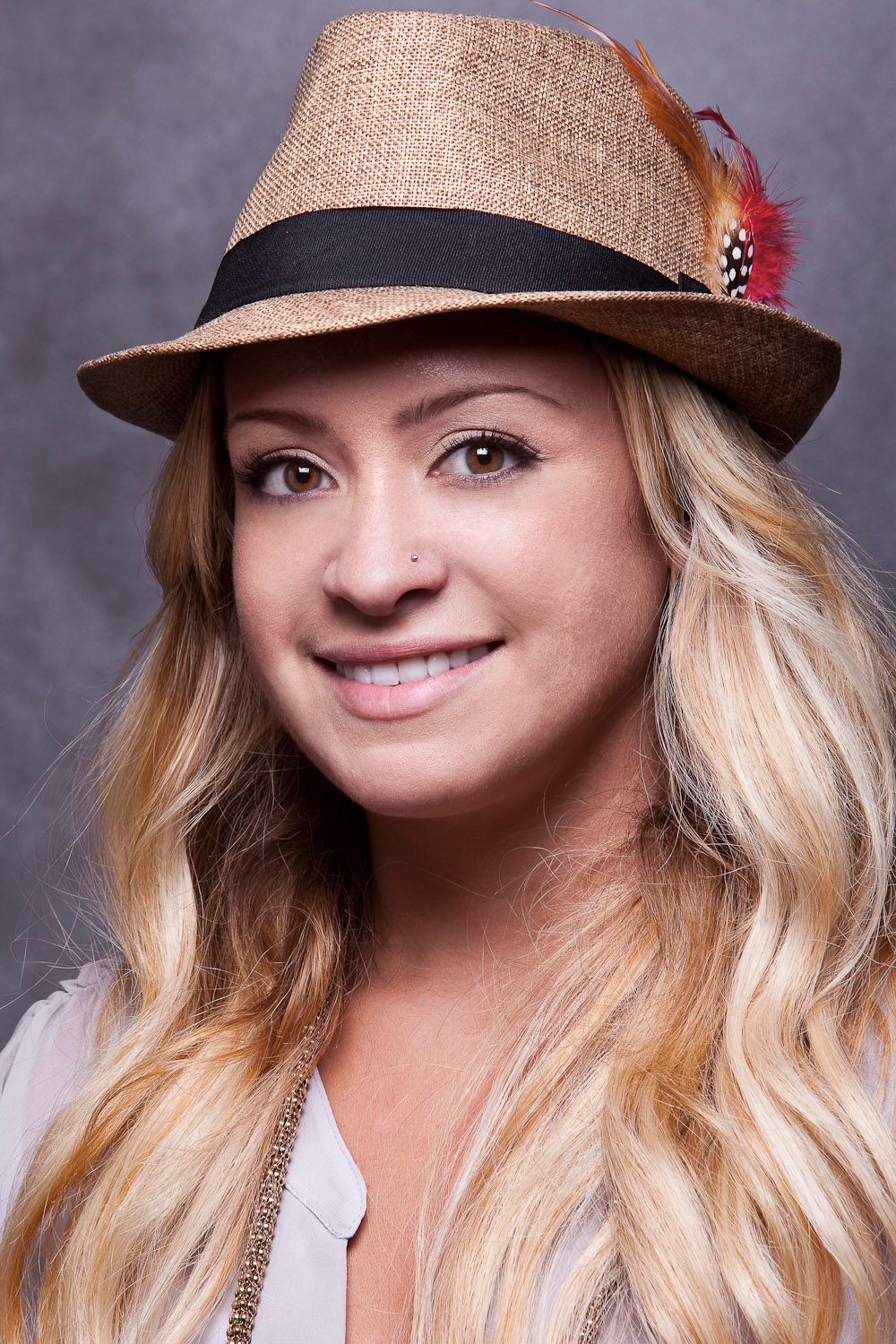 Smiling blonde woman wearing a straw fedora hat with feather accent in a studio portrait.