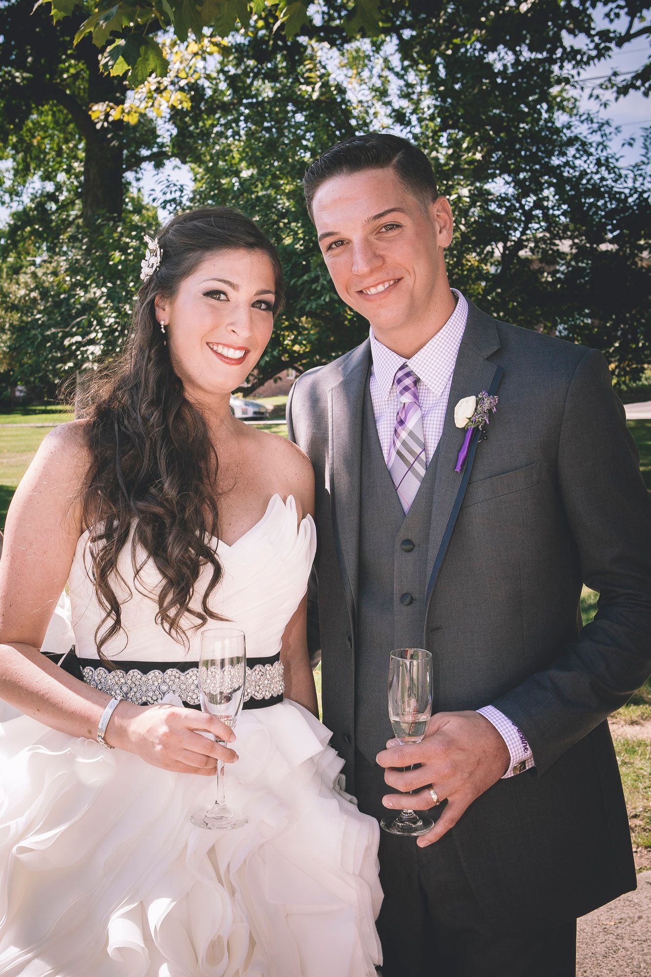 Smiling bride and groom hold champagne glasses outdoors under green trees after wedding ceremony.