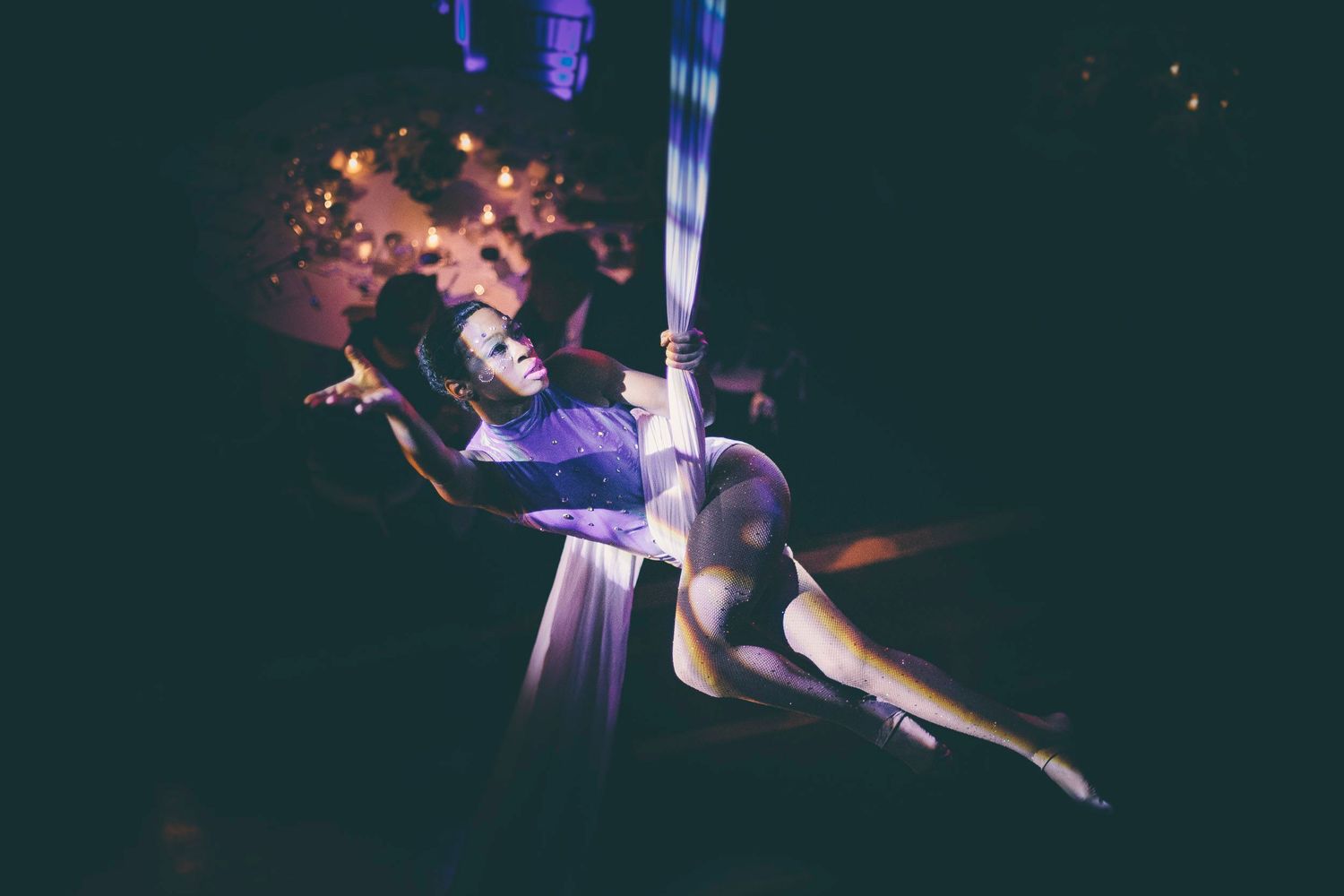 An aerial performer reclines gracefully in white silk fabric under warm purple event lighting.