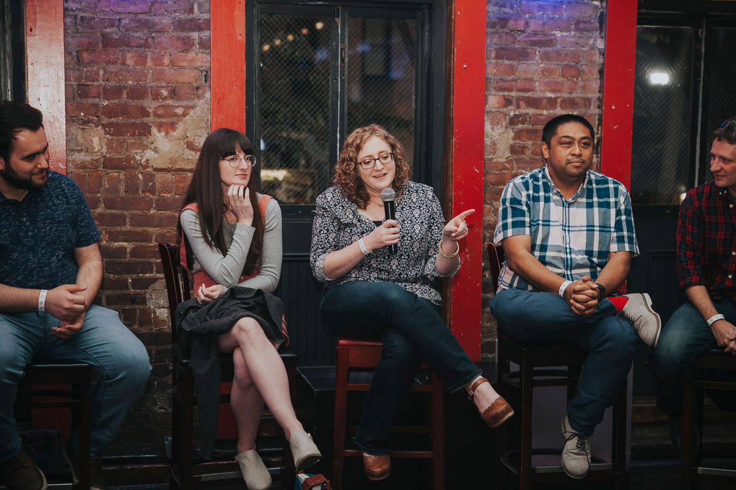Five panelists seated on stools during a discussion event in a brick-walled venue.