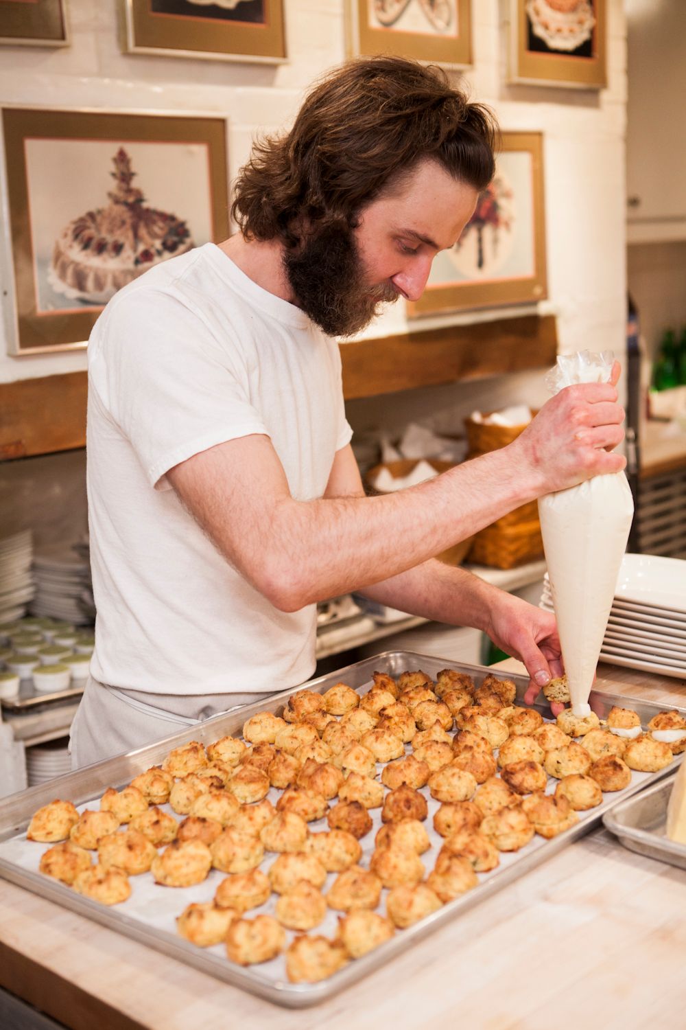 Bearded chef using a piping bag to fill golden baked gougeres on a baking sheet.
