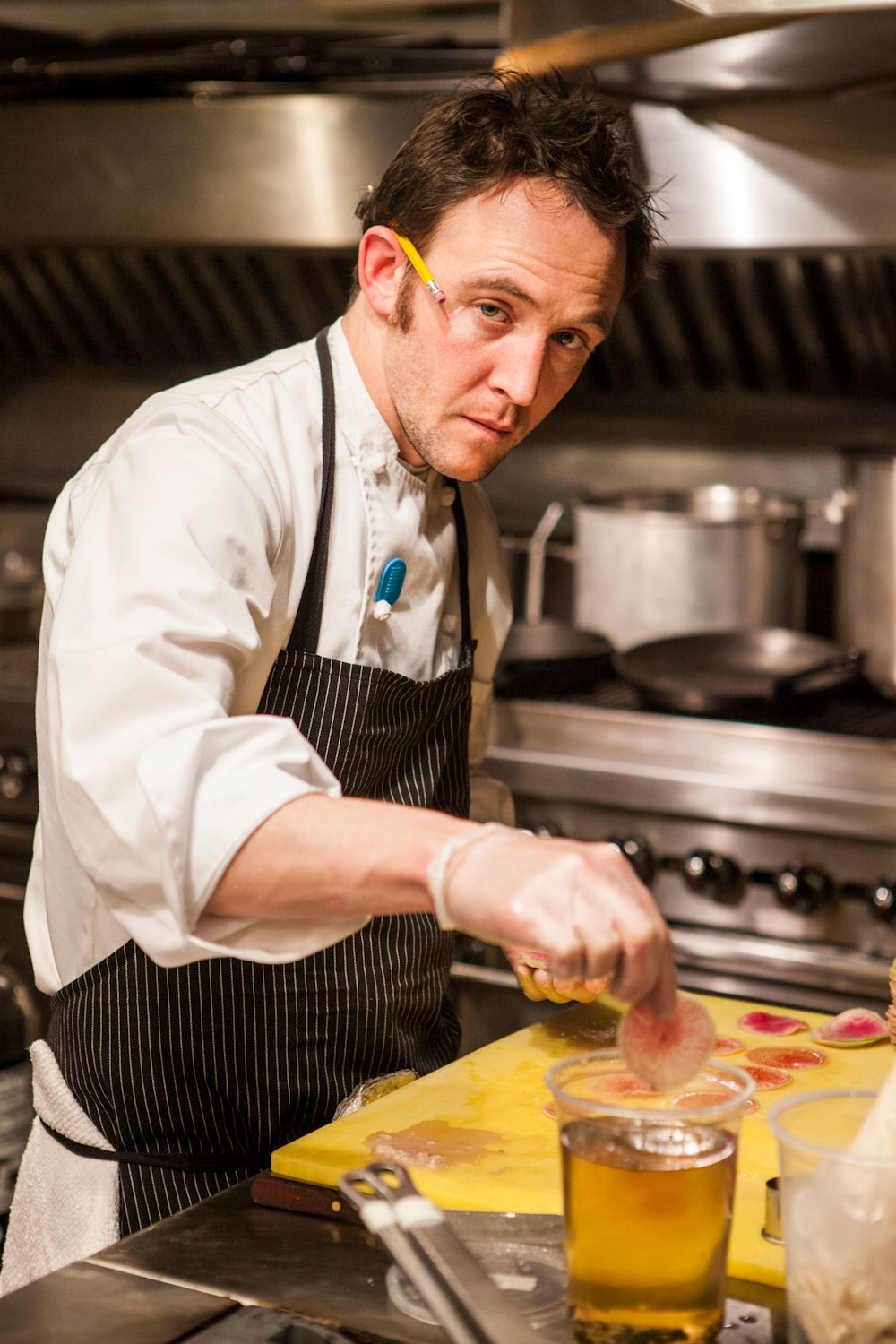 Male chef in striped apron working with ingredients on a yellow cutting board in a restaurant kitchen.