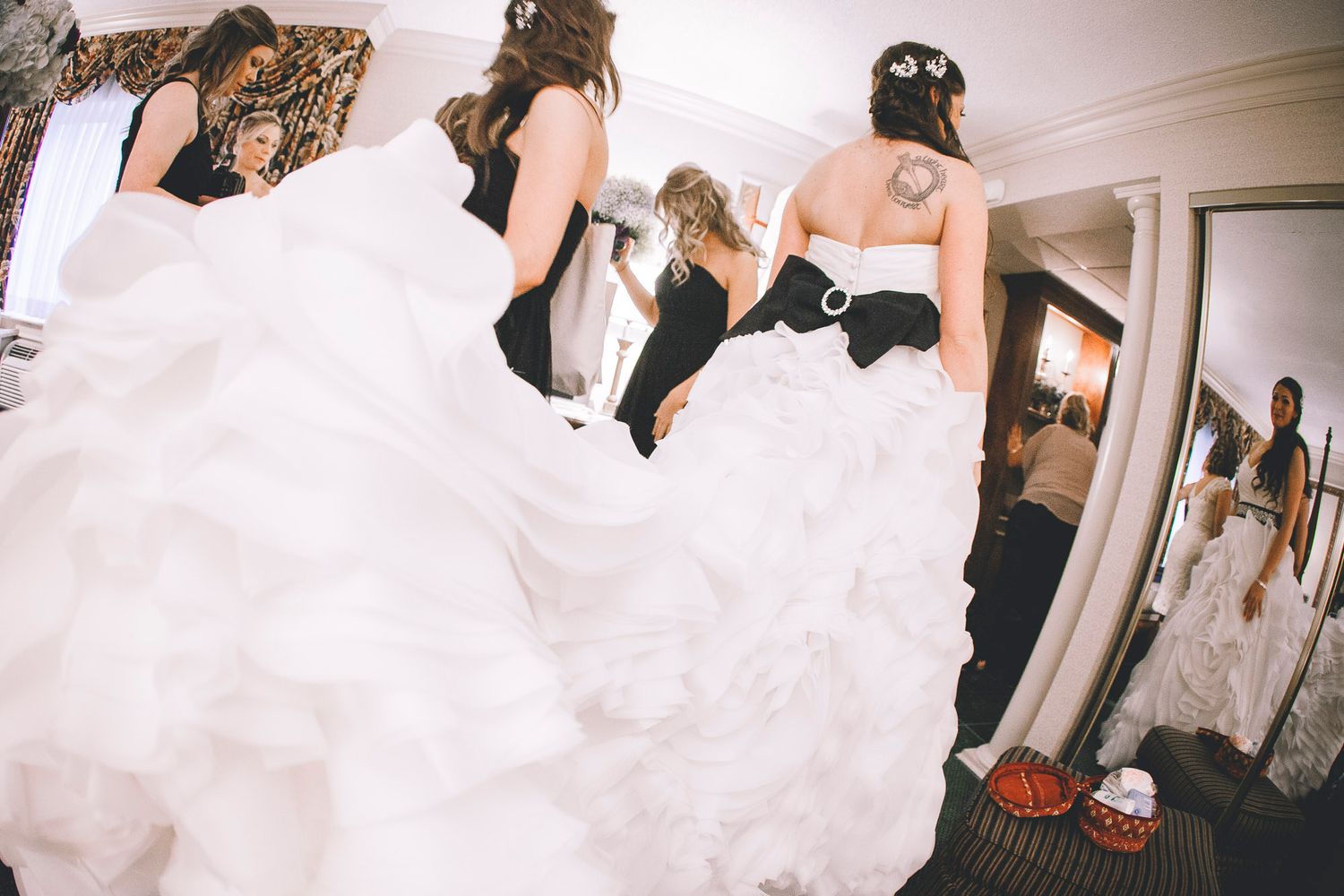 Bridesmaids in black dresses helping adjust the voluminous white ruffled wedding gown of the bride indoors.