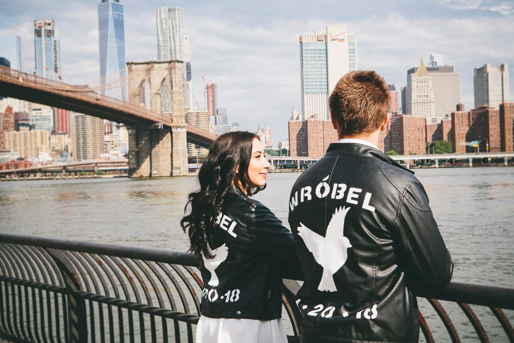 Couple in matching custom black leather jackets standing by railing with Brooklyn Bridge and NYC skyline behind them.