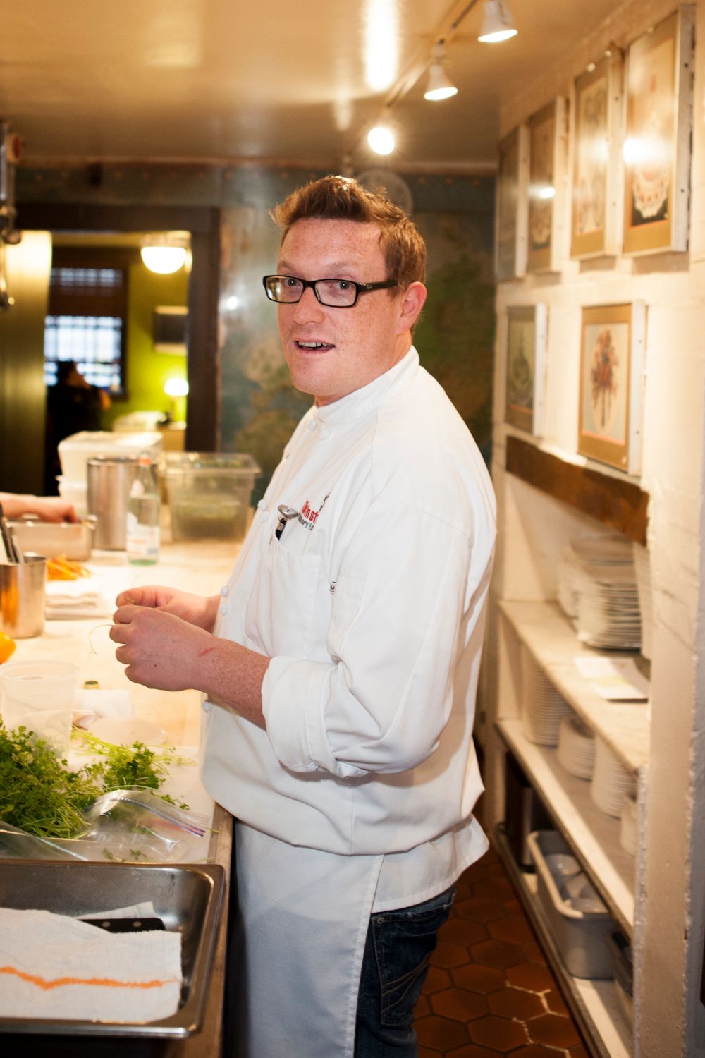 Smiling chef in white jacket and glasses standing at a kitchen prep station with fresh herbs.