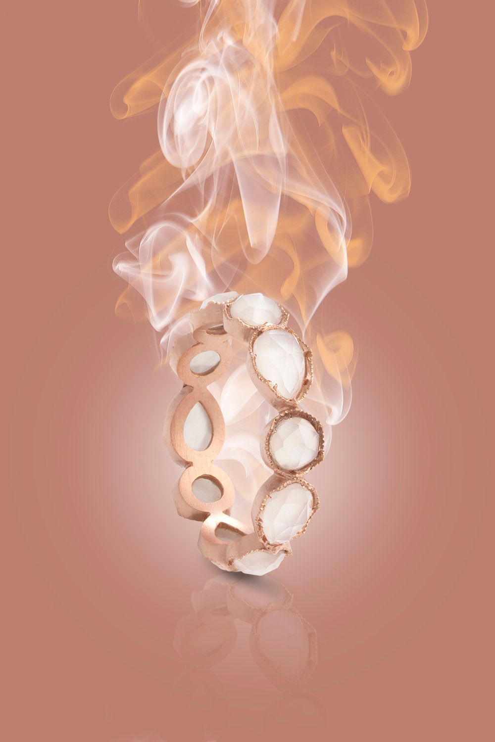 Rose gold open-work bracelet with white oval stones floating in smoke on pink background.