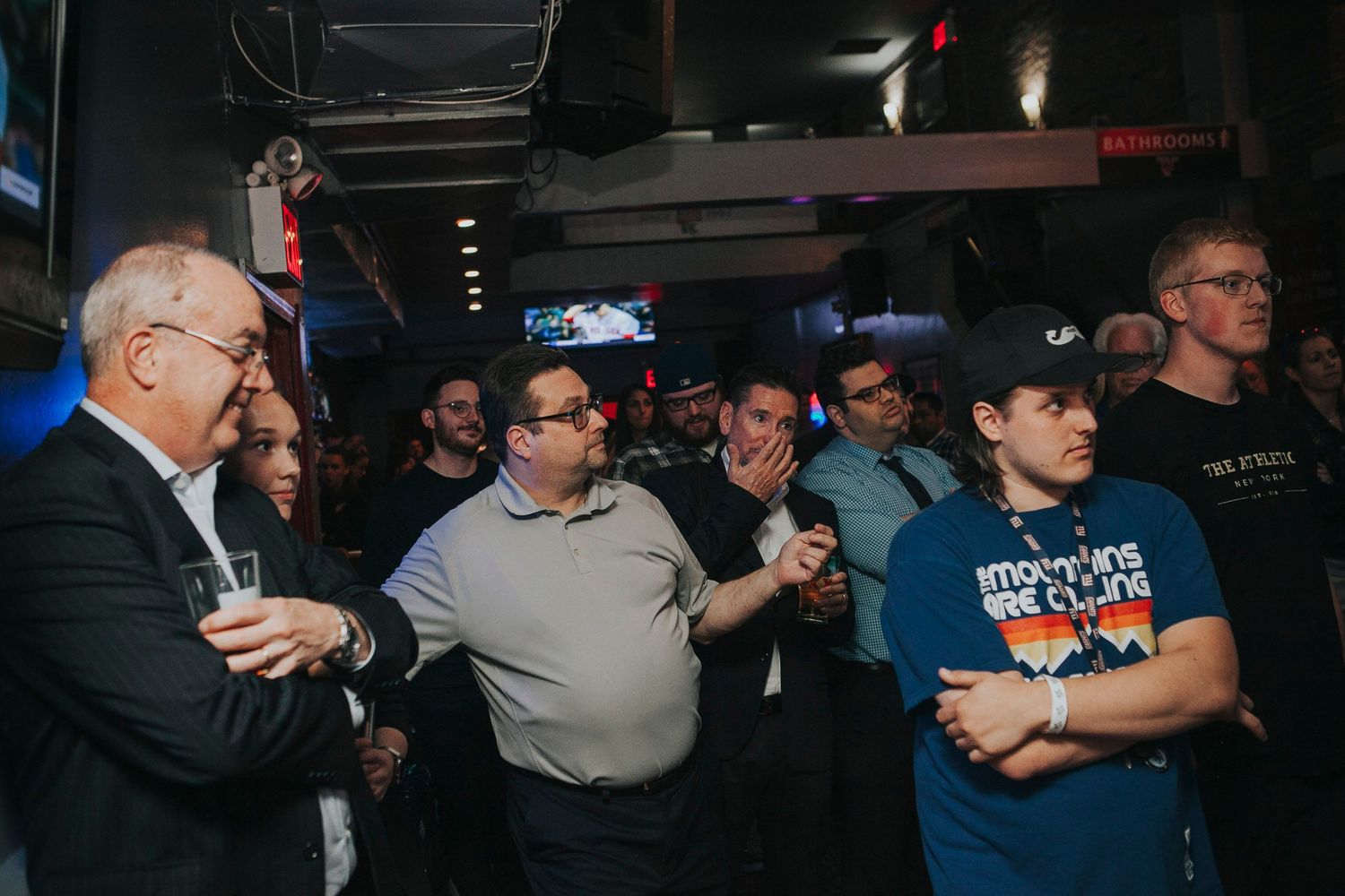 Group of people gathered in a dimly lit bar watching an event on a screen.