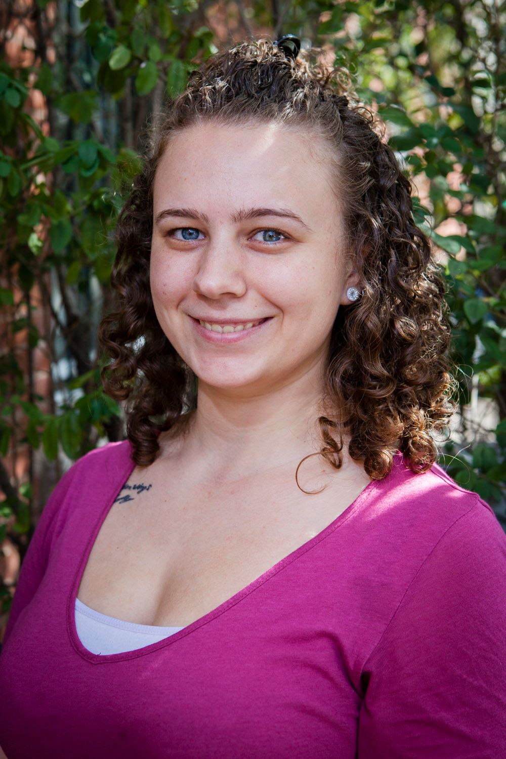 Young woman with curly hair and blue eyes smiling in a pink top outdoors.