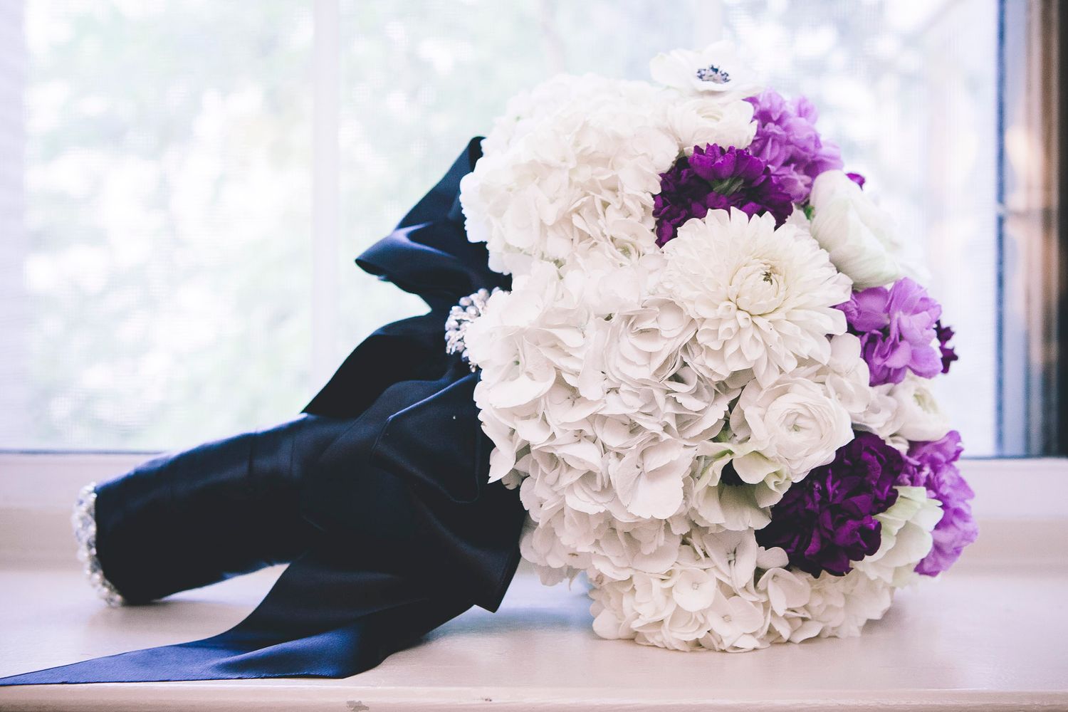 Elegant wedding bouquet with white hydrangeas, purple flowers, and a navy blue satin ribbon on a windowsill.