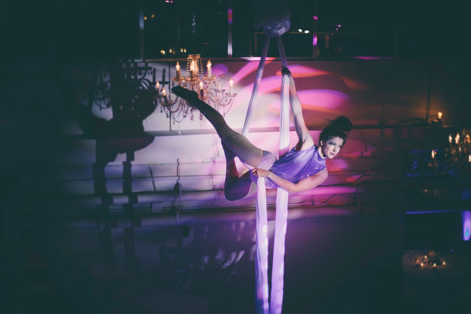 Aerialist performing on hanging silks in a candlelit ballroom with purple ambient lighting.