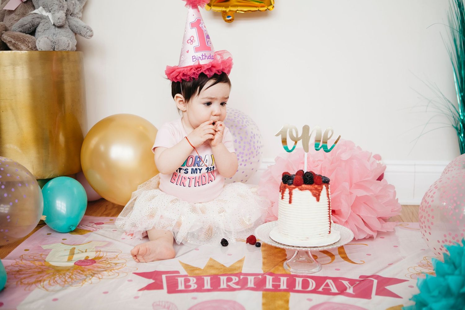 Baby girl in pink party hat and tutu eats cake during her first birthday cake smash celebration.
