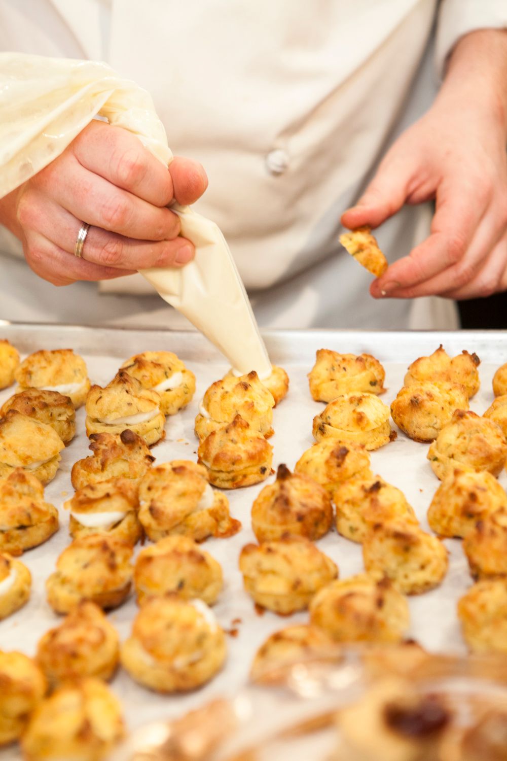 Close-up of hands using a piping bag to fill freshly baked cheese puffs on parchment paper.