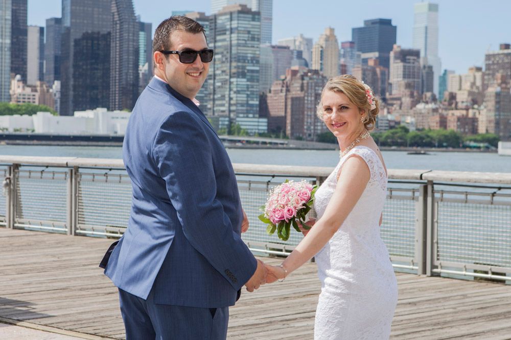 Newlyweds hold hands on waterfront pier with NYC skyline backdrop, bride holding pink rose bouquet.