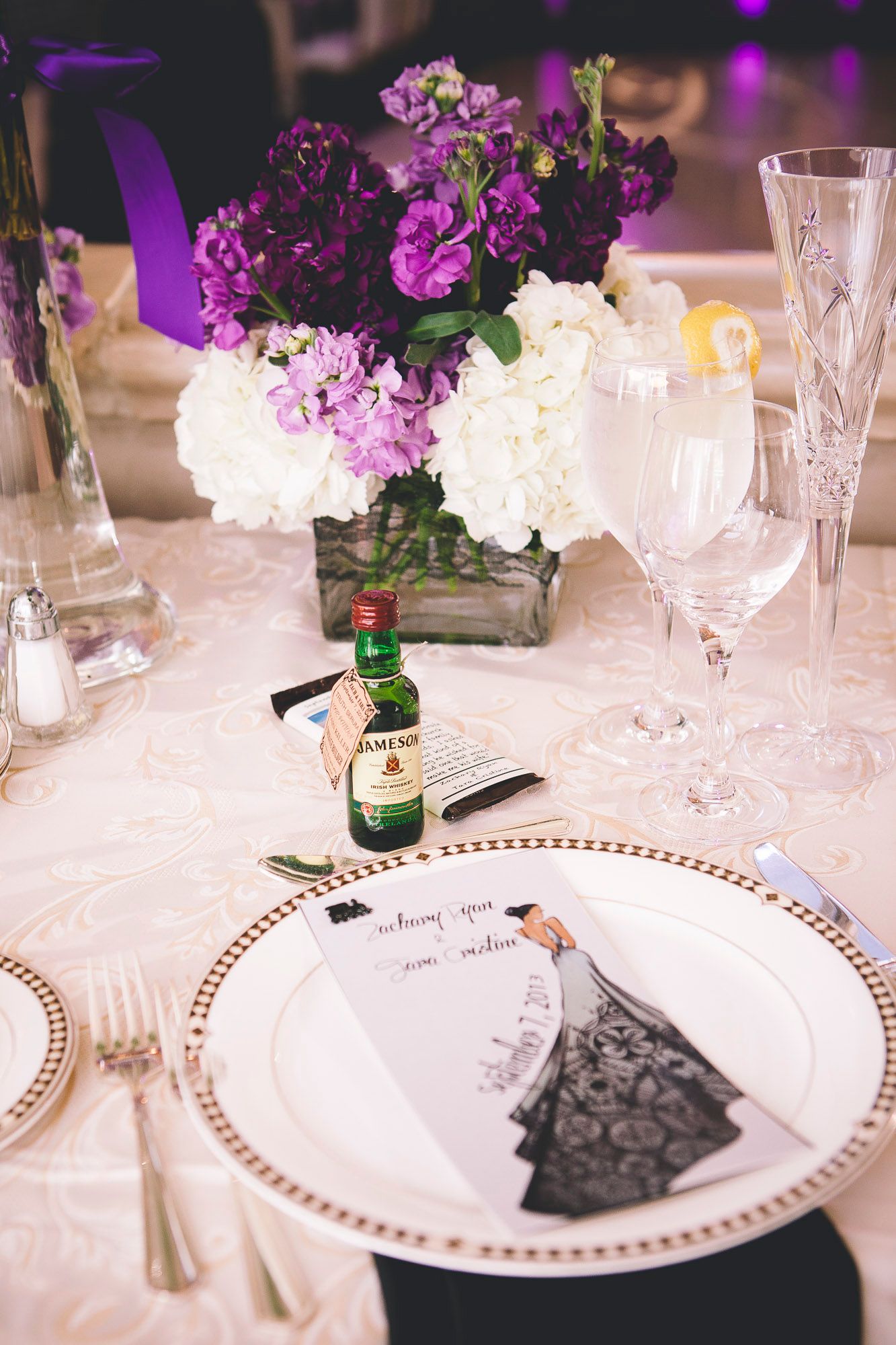 Elegant wedding reception table setting with purple floral centerpiece, crystal glassware, and a mini Jameson whiskey bottle.