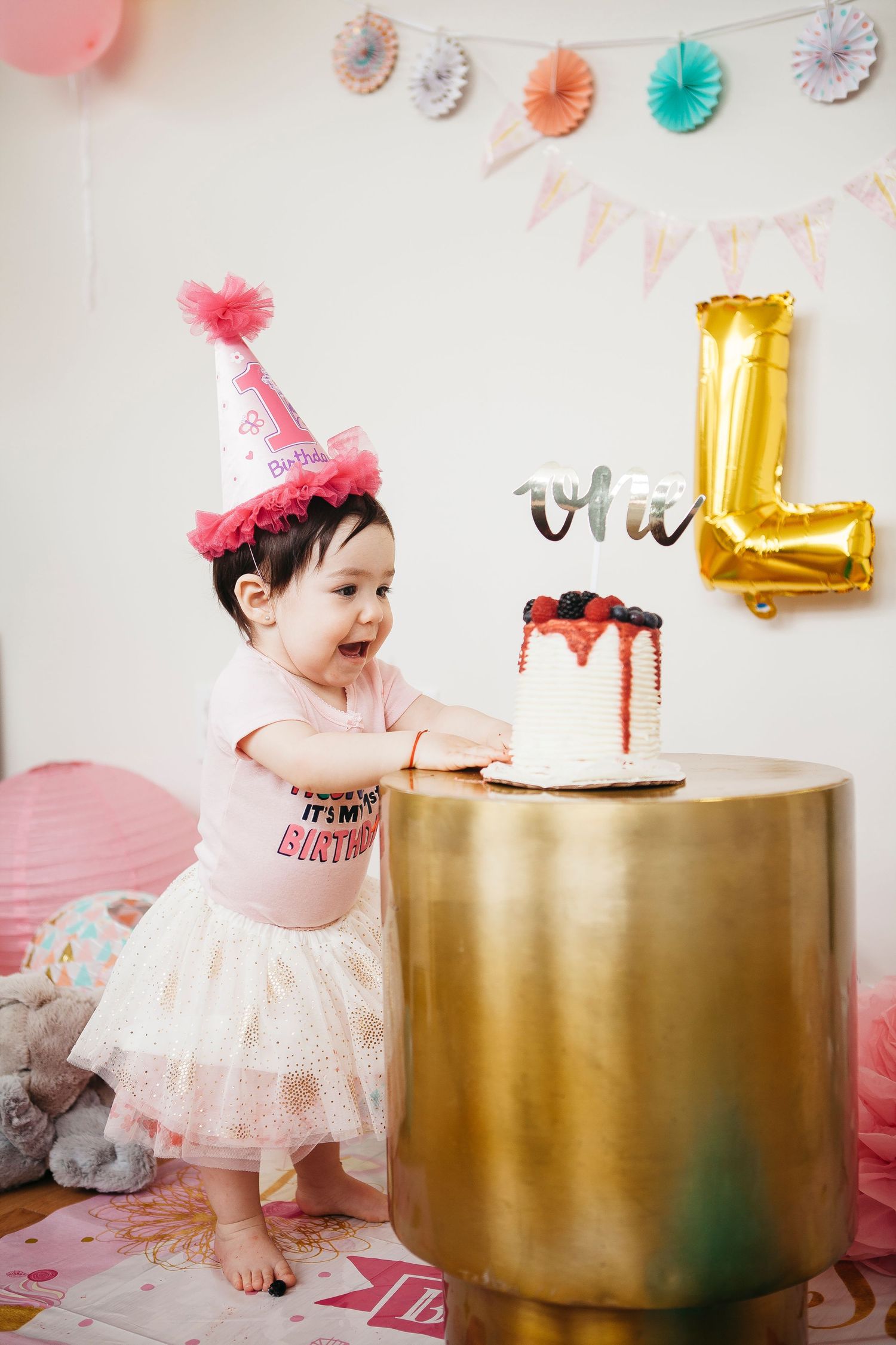 Excited baby girl in pink birthday hat stands at gold table reaching for her first birthday cake.